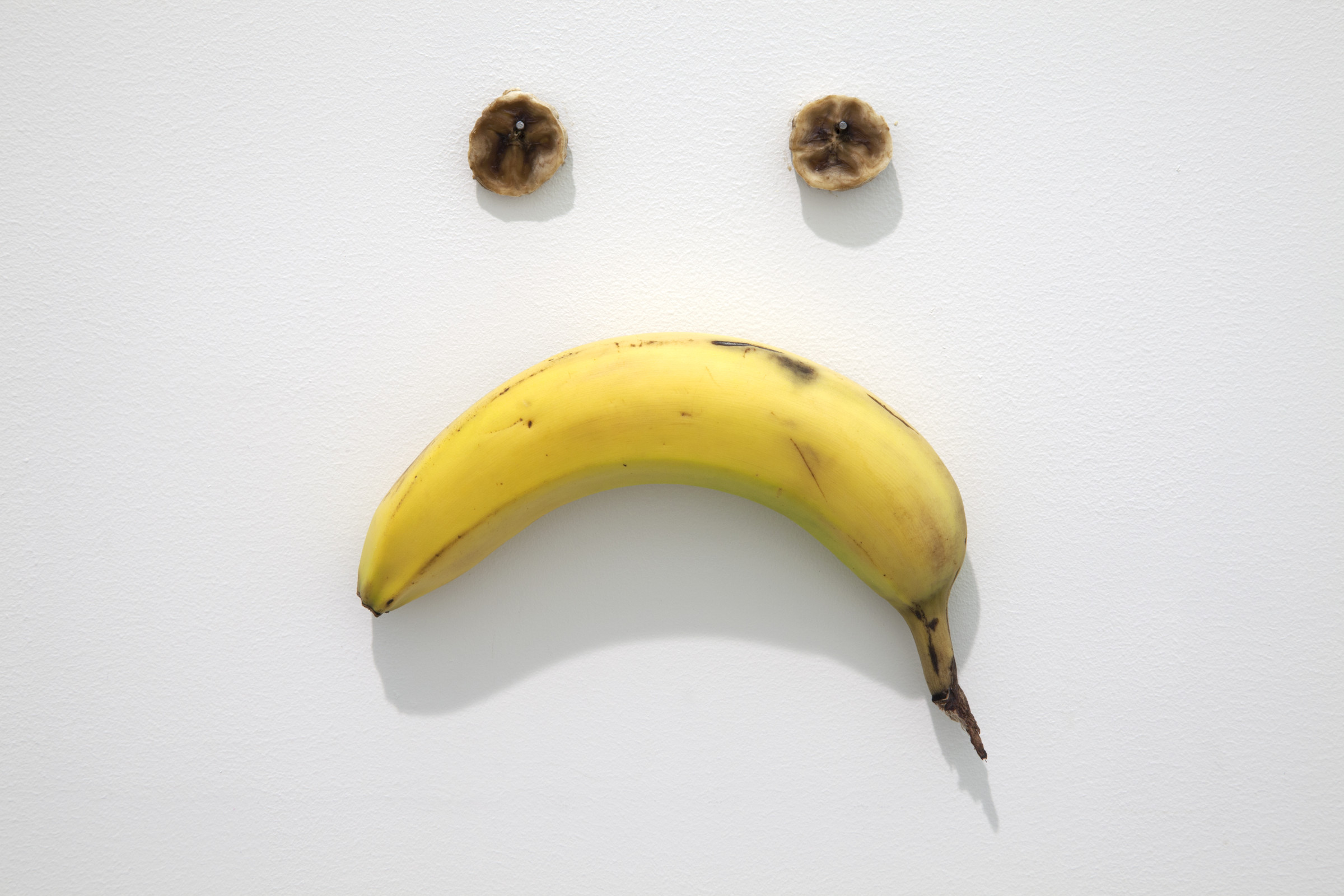 A close-up image of a banana pinned horizontally to a wall with the bend pointed down to form a frown, with two slices of banana pinned above it to form eyes
