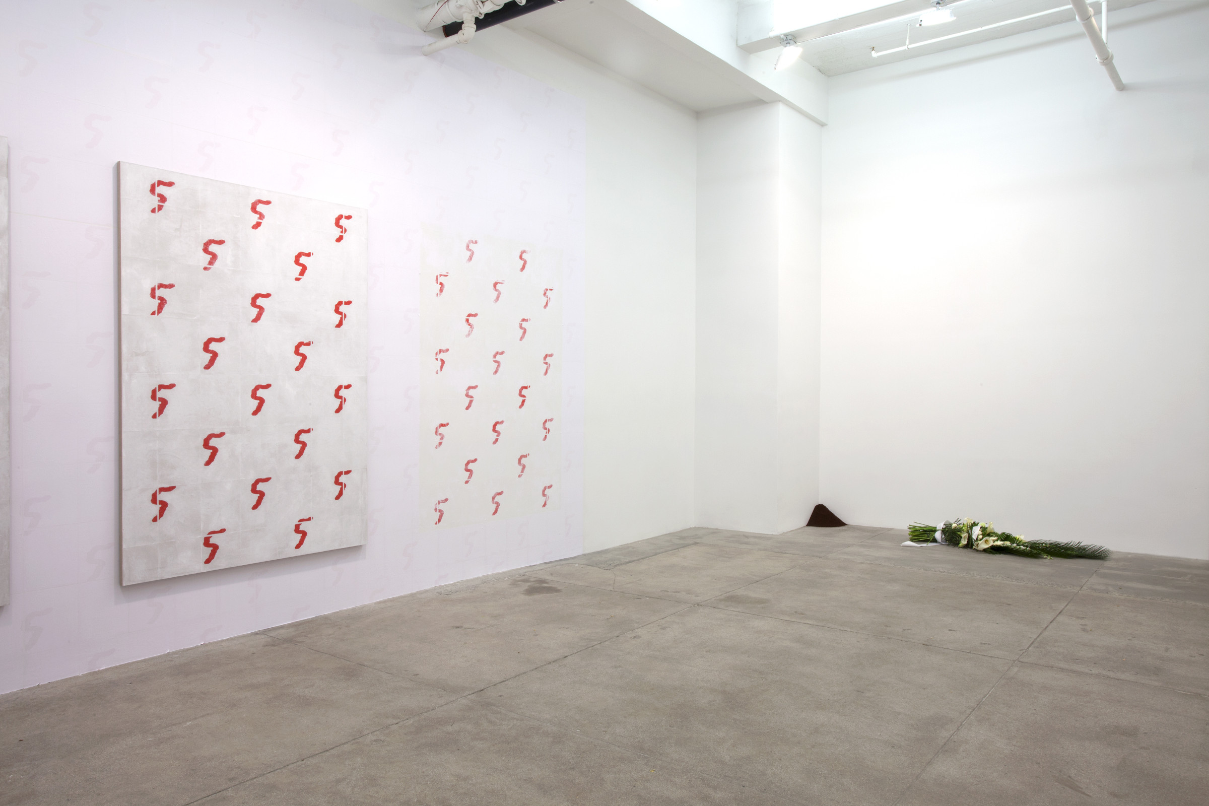 An installation view of an exhibition showing a painting patterned with a red icon like a reversed question mark, a wallpaper rubbed away to reveal a similar pattern, a pile of coffeee grinds in the corner and an elaborate bouquet laid on its side on the floor