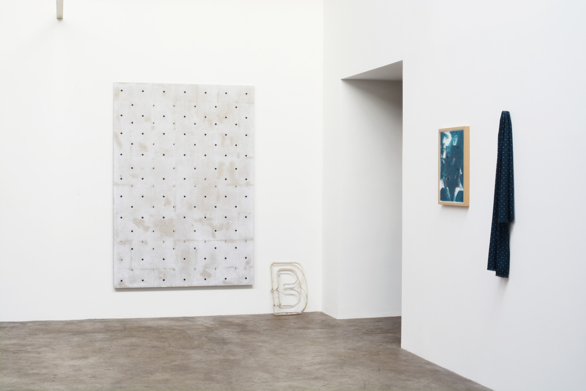 And installation view of an exhibition featuring multiple works and objects in a corner: a large polka-dot painting on the wall, a set of old, white, neon letters (a D and a B), neither connected nor on, a framed work with a blue screen print of a face on the glazing, and a polka-dot scarf hanging on the wall
