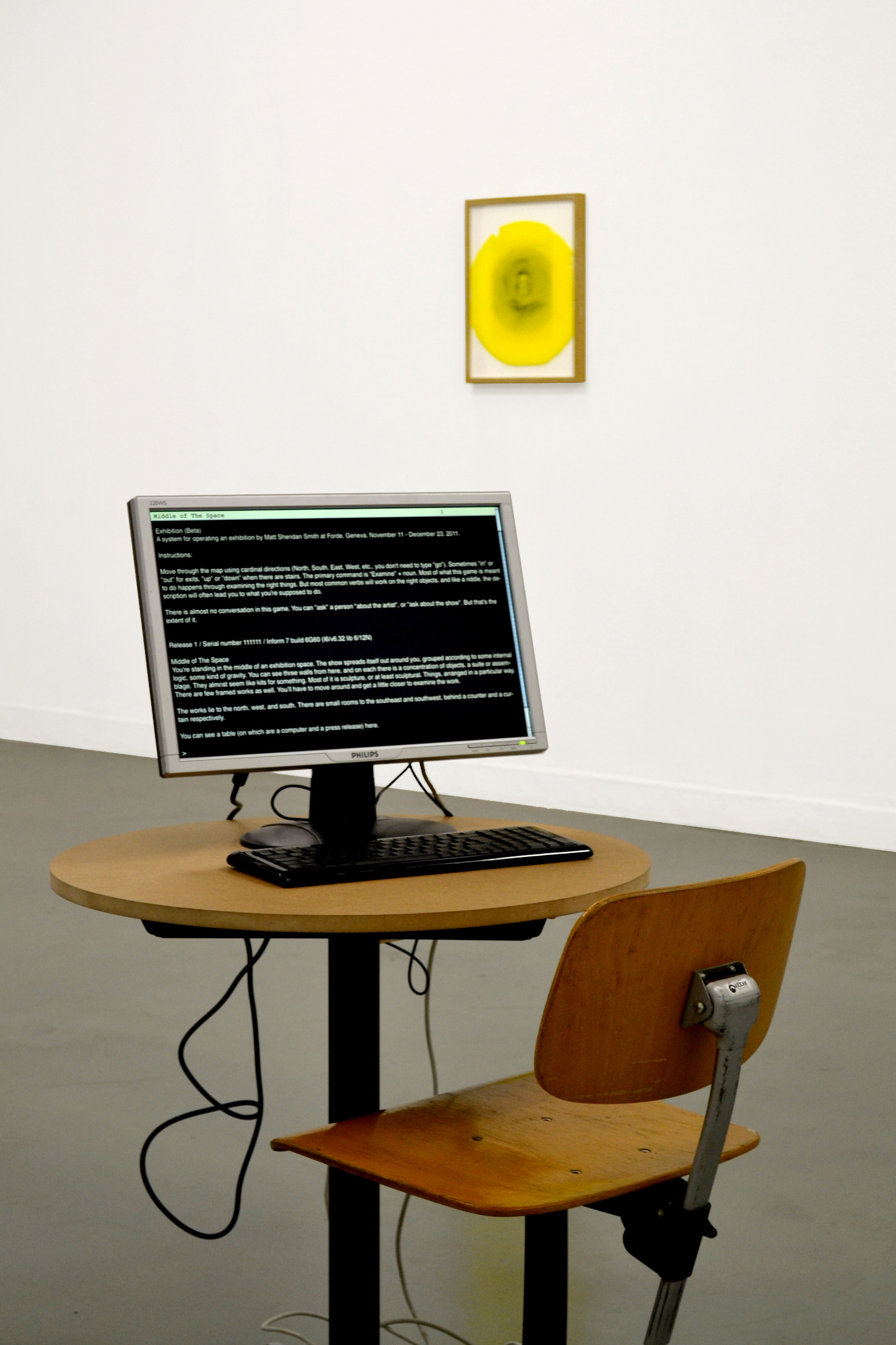 A n installation view of an exhibition. In the foreground is a small round table and a wooden school chair. On the table is a computer monitor and a keyboard. On the screen is a text-based adventure game. It is impossible to read from the image but shows white text on a black background. In the background is a black curtain to the left and a framed work with a yellow, inky print of a woman's face, printed diretly on the glazing.