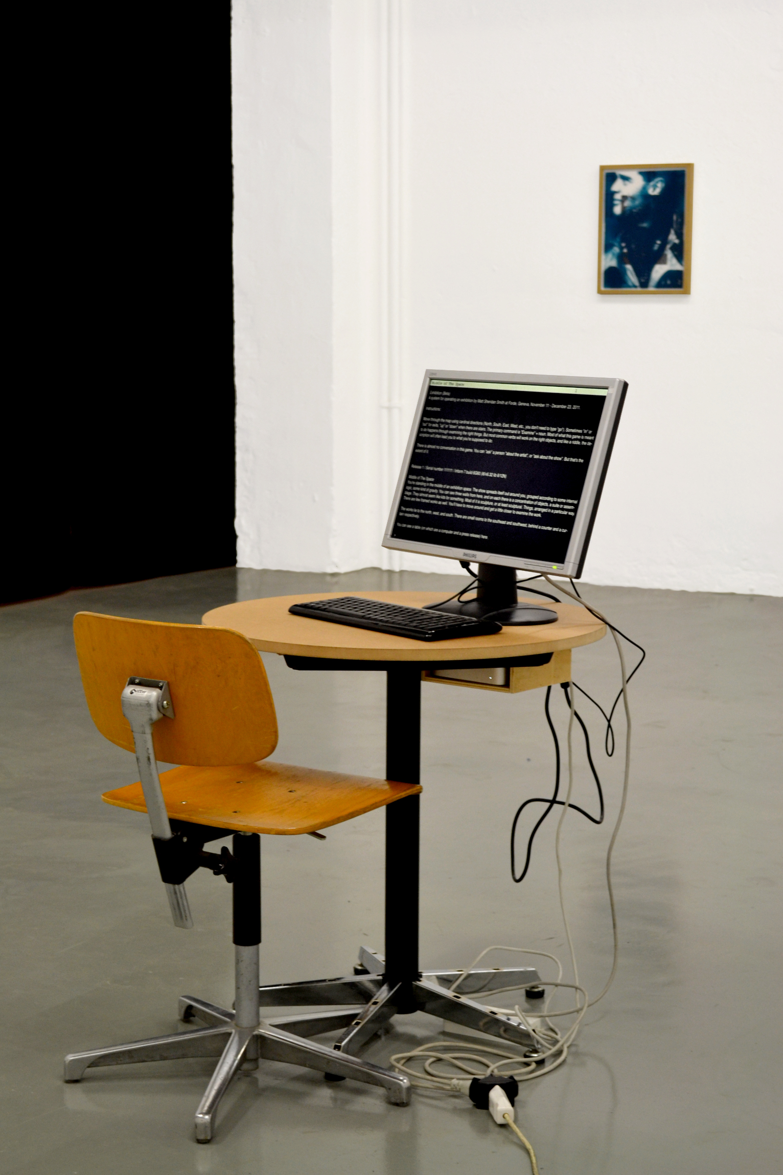 A n installation view of an exhibition. In the foreground is a small round table and a wooden school chair. On the table is a computer monitor and a keyboard. On the screen is a text-based adventure game. It is impossible to read from the image but shows white text on a black background. In the background is a framed work with a dark blue, inky print of a mans face looking to the right printed diretly on the glazing.