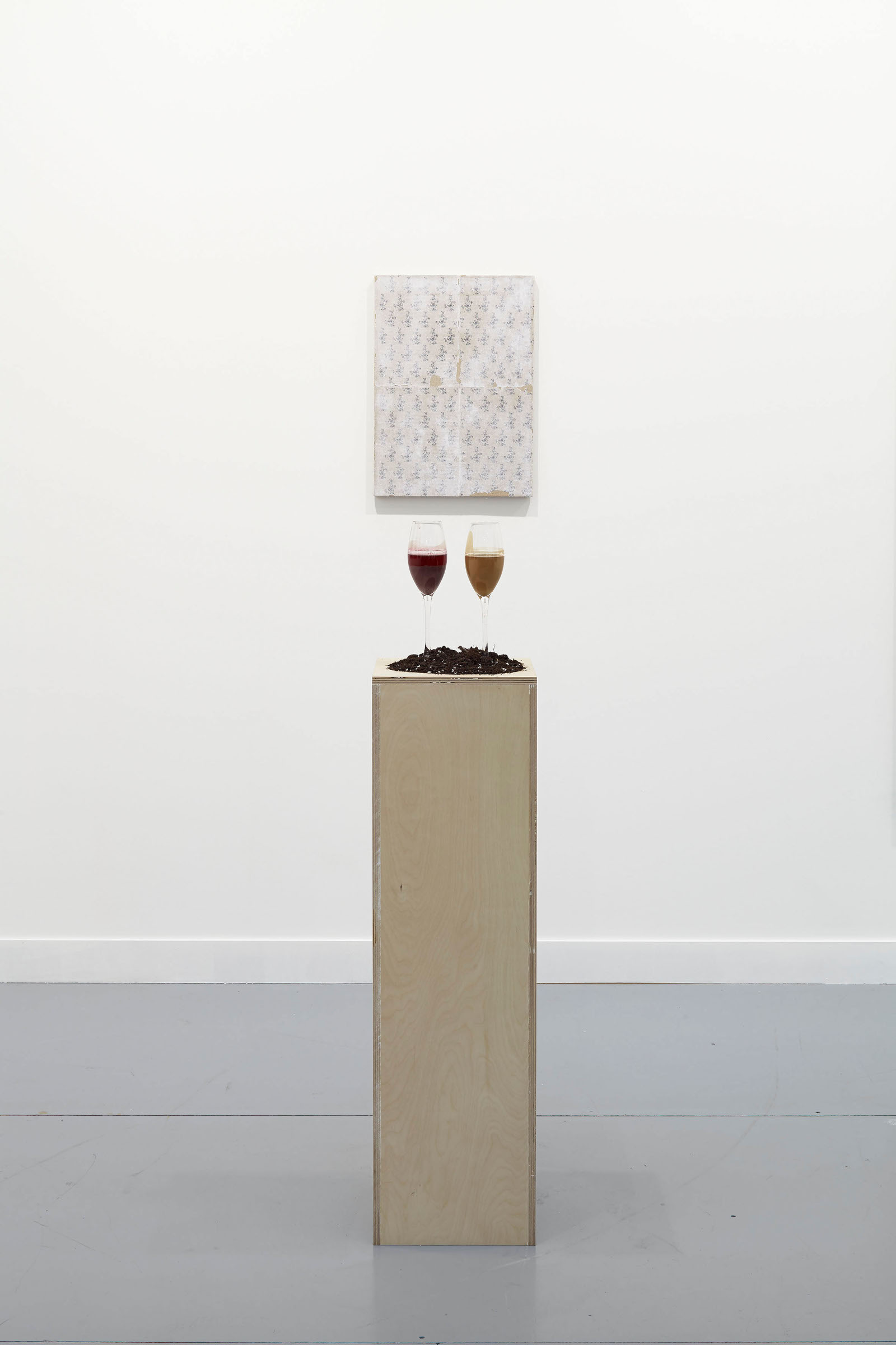 A straight on image of a plywood pedestal about 10 inches by 10 inches by 45 inches. On the pedestal: a small pile of soil and two champagne flutes with thick, viscous fluids that are the byproduct of champagne production. One is dark red and one is light brown. In the background is a small patterned painting.