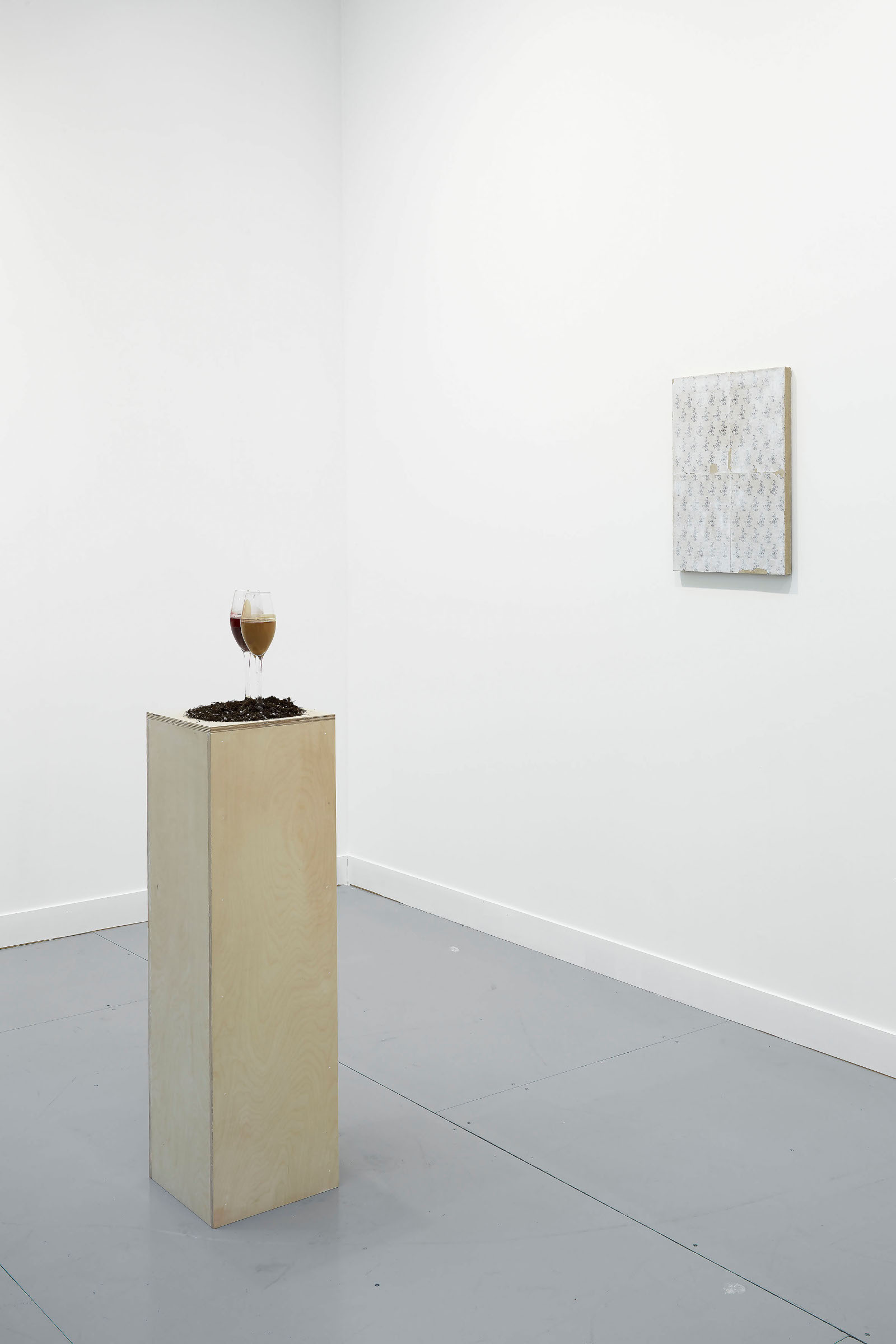 An image shot from a 45 degree angle of a plywood pedestal about 10 inches by 10 inches by 45 inches. On the pedestal: a small pile of soil and two champagne flutes with thick, viscous fluids that are the byproduct of champagne production. One is dark red and one is light brown. In the background is a small patterned painting.
